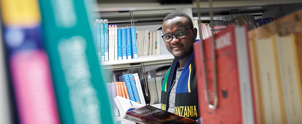 A man holding an open book, wearing glasses, and a scarf of the Tanzanian flag standing in-between library book shelves